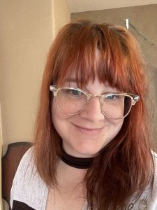 The author, a white femme with red hair, bangs, large glasses, and a smirk.